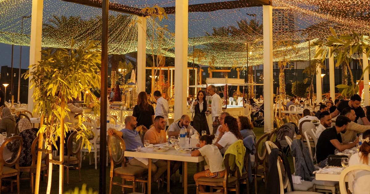People sitting at tables under an open air Iftar tent with lights