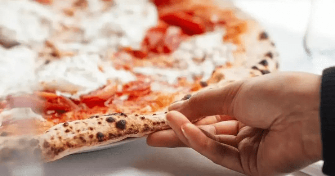 Close-up of a hand gently holding a slice of pizza at L'Antica Pizzeria, Dubai Hills Mall. The pizza features a thin, crispy crust and is topped with a vibrant tomato sauce and fresh mozzarella.