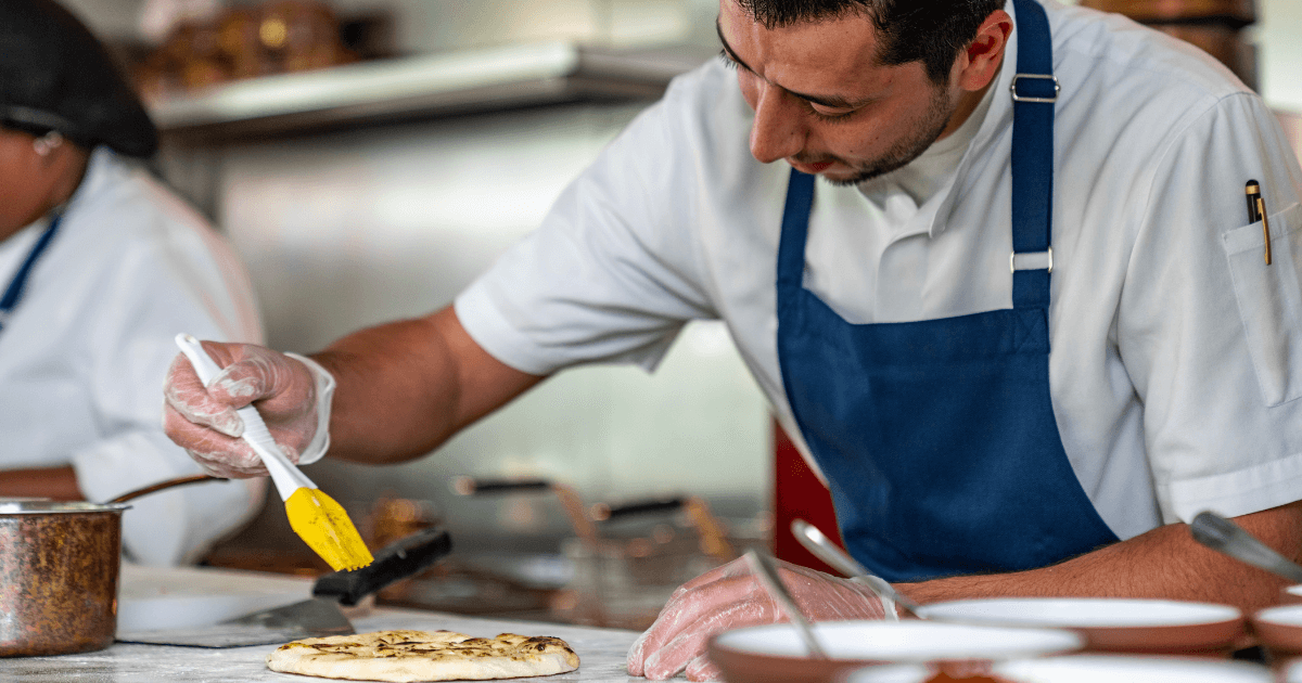 A focused chef in a kitchen at Assembly restaurant, Expo City Dubai, garnishes a flatbread, showcasing culinary expertise and attention to detail in a professional restaurant setting.