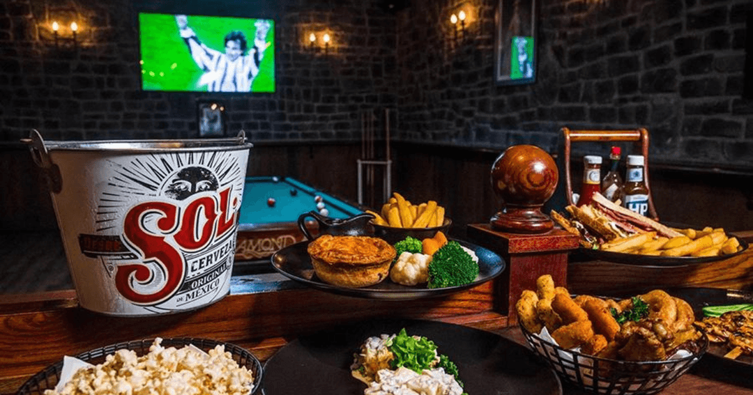 A vibrant sports bar setup at Nell Gwynne, featuring a bucket of Sol beer and a variety of pub foods like fries, pie, and chicken wings. A pool table and a large TV screen showing a soccer match create a lively and engaging environment.